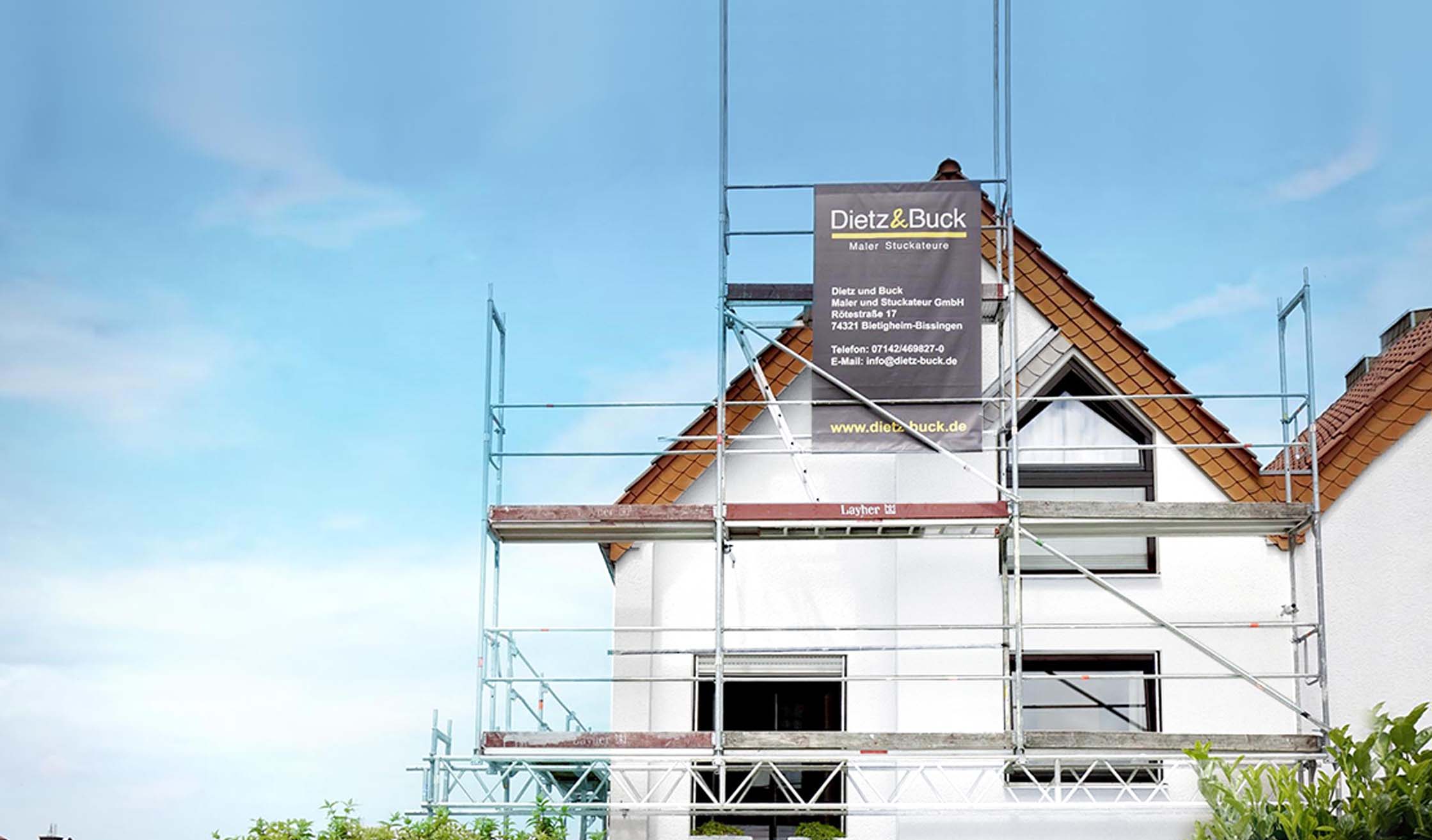 Bild vom Baugerüst mit einem Banner von Dietz und Buck vor einem weißen Haus für Fassadenrenovierung.