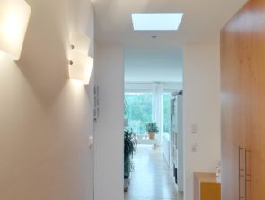 Bild von einem Flur ins Wohnzimmer mit weißen Wänden und Holzmöbeln nach einer Wohnungsrenovierung, von Tageslicht und Wandleuchten beleuchtet.